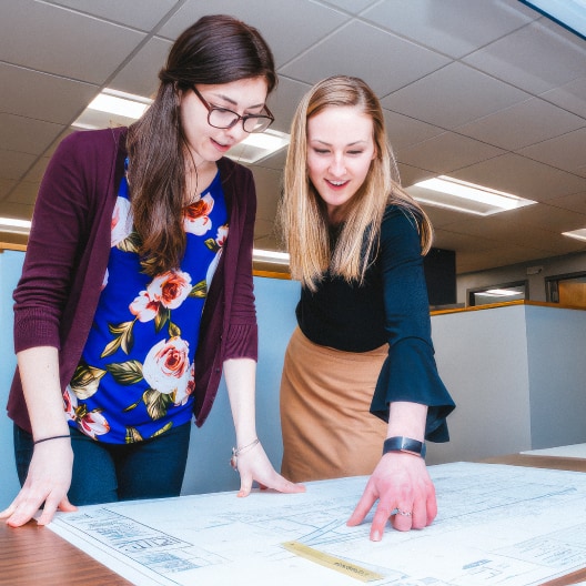 Two women stand at a desk, examining large architectural blueprints. One woman points at a specific spot on the plans while the other looks on. Both appear focused and engaged in discussion.