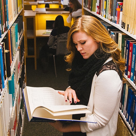 A woman with long red hair reads a large book between bookshelves in a library, while two other people sit at desks in the background.