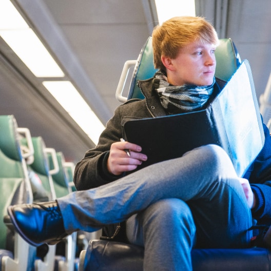 A person with light hair sits on a train seat, legs crossed, wearing a coat and scarf, and holding an open black folder. The train cabin appears modern and well-lit.