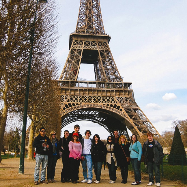 A group of people poses for a photo in front of the Eiffel Tower in Paris on a clear day, with trees lining the path beside them.