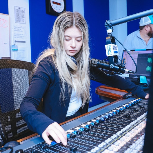 A woman with long blonde hair sits at a radio station soundboard, adjusting controls. She wears a dark jacket and headphones around her neck. A microphone, computer monitor, and another person are visible in the background.