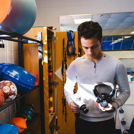 A person in a gray shirt examines a black kettlebell and a white water bottle in a gym, surrounded by colorful exercise equipment and sports balls.