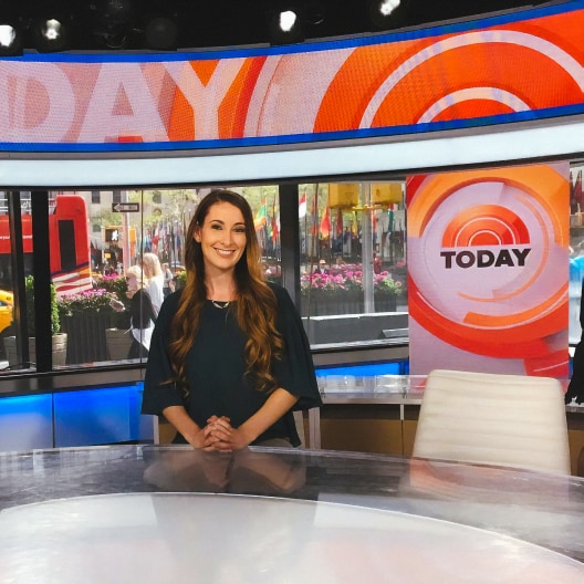 A woman with long brown hair smiles while sitting at a news desk on the set of the Today Show, with bright studio lights and a city street visible through large windows behind her.