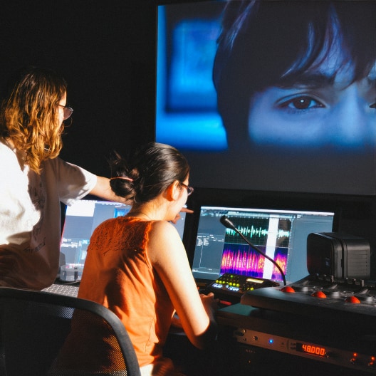 Two people work in a dimly lit studio editing video and audio on computer screens, while a large projected image of a person’s face appears on the wall in front of them.