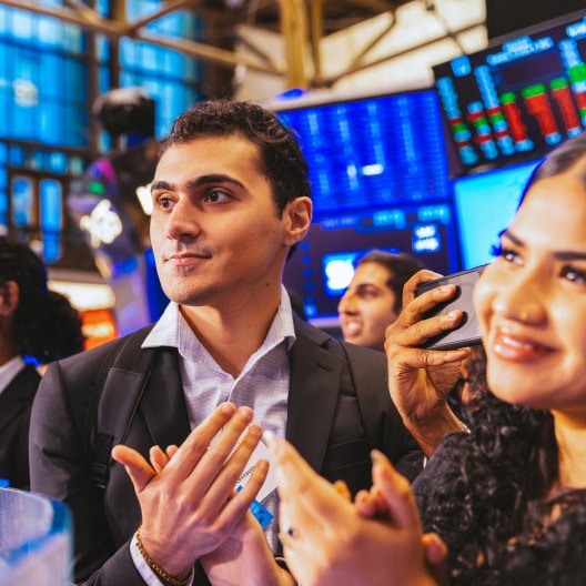 A group of professionally dressed people stand closely together, clapping and smiling, in front of large digital screens displaying stock market data in a busy trading environment.
