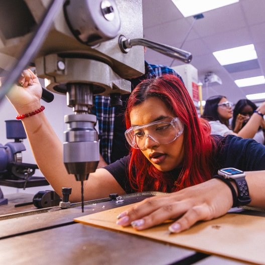 A young woman with red hair and safety goggles operates a drill press machine in a workshop, focusing on a piece of wood while others work in the background.