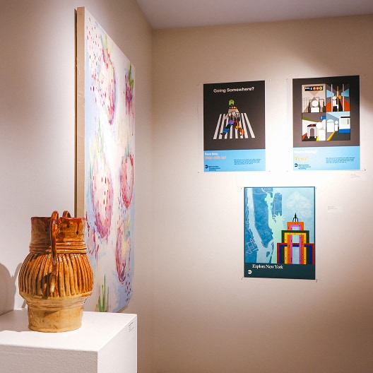 A ceramic vase sits on a white pedestal near a wall displaying colorful abstract art, including a large canvas and three vibrant posters with graphic designs and cityscapes.