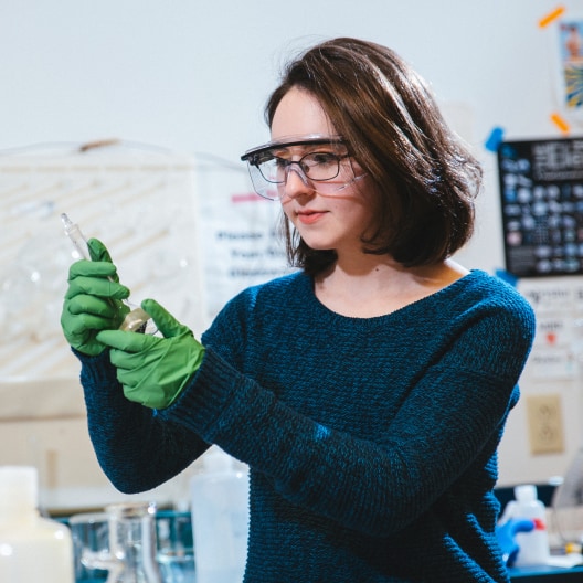A woman wearing safety goggles and green gloves is using a dropper to transfer liquid into a small container in a laboratory setting.