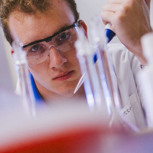 A young scientist wearing safety goggles and a lab coat uses a pipette in a laboratory, concentrating on transferring liquid between glass containers.
