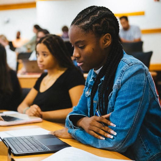 Two students sit in a classroom, focused on their laptops. The student in the foreground wears a denim jacket and has braided hair. Other students and desks are visible in the blurred background.