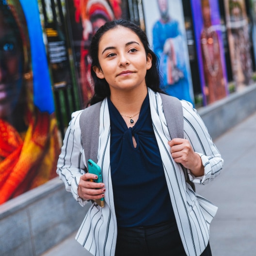 A young woman with a backpack stands on a city sidewalk, holding a phone and looking thoughtful. Colorful posters with faces are displayed on the wall behind her.