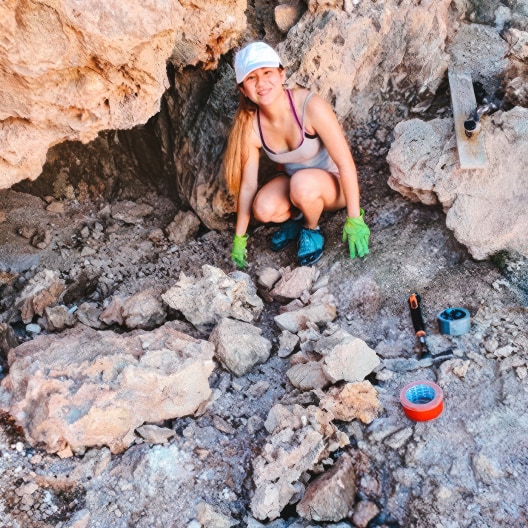 A woman wearing a white cap, tank top, shorts, green gloves, and blue shoes kneels on rocky ground at a dig site. Excavation tools and tape are scattered nearby. She is smiling and appears to be working outdoors.