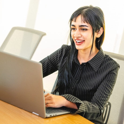 A woman with long dark hair, wearing a black pinstripe shirt, sits at a desk smiling while using a laptop in a bright office setting.