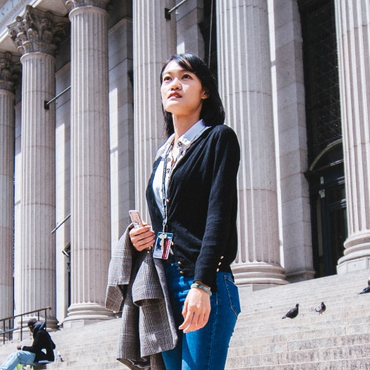 A woman stands on outdoor steps in front of a large building with tall columns, holding a coat and phone. She looks upward with a thoughtful expression.