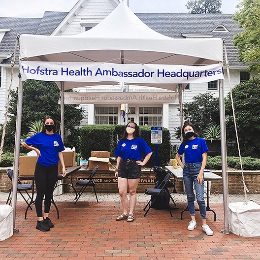 Three people wearing masks and blue shirts stand under a white tent labeled Hofstra Health Ambassador Headquarters, with tables and boxes behind them, in front of a white building.