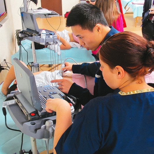Two healthcare professionals examine a printout and monitor on medical equipment, likely performing or analyzing an ultrasound exam in a clinical setting. Other patients and staff are visible in the background.