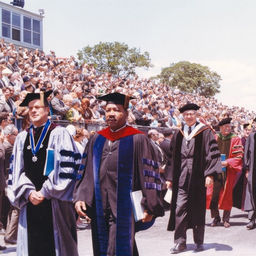 A group of faculty in academic regalia walk in a graduation procession, with a large crowd of spectators seated in bleachers under a sunny sky in the background.