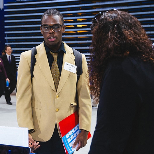 A young man wearing glasses, a beige blazer, and a tie speaks to a woman at what appears to be a networking event or career fair. He holds a red folder and wears a name tag. Other people are visible in the background.