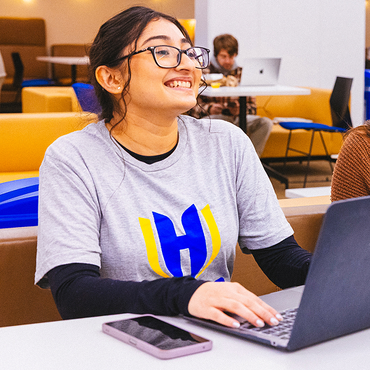 A smiling person with glasses types on a laptop in a brightly lit room. They wear a gray T-shirt with a large blue and yellow H design. A phone is on the table nearby, and others work in the background.