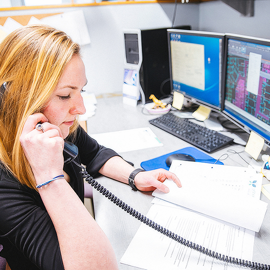 A woman with blonde hair talks on a landline phone while working at a cluttered desk with papers and two computer monitors displaying maps and documents.