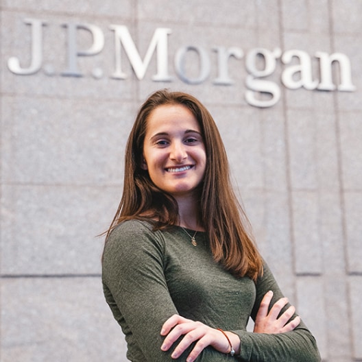 A person with long brown hair, wearing a green long-sleeve top, stands smiling with arms crossed in front of a gray wall displaying the J.P. Morgan logo.