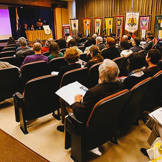 A group of people sits in an auditorium facing a stage where speakers are presenting. Colorful banners with various symbols and crests line the back of the stage and room, giving a formal atmosphere to the event.