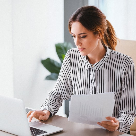 A woman in a striped shirt sits at a desk, looking at a laptop screen while holding documents in her hand. A green plant is visible in the background.