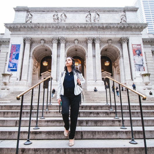 A woman in business attire walks down the steps outside a grand, historic building with columns and arched entrances, likely a library or museum, with banners and people in the background.