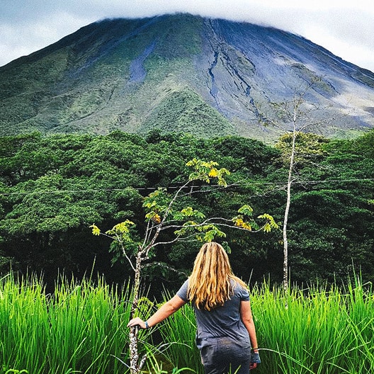A woman with long blonde hair stands in tall green grass, facing a lush forest and a large, tree-covered volcano under a cloudy sky.