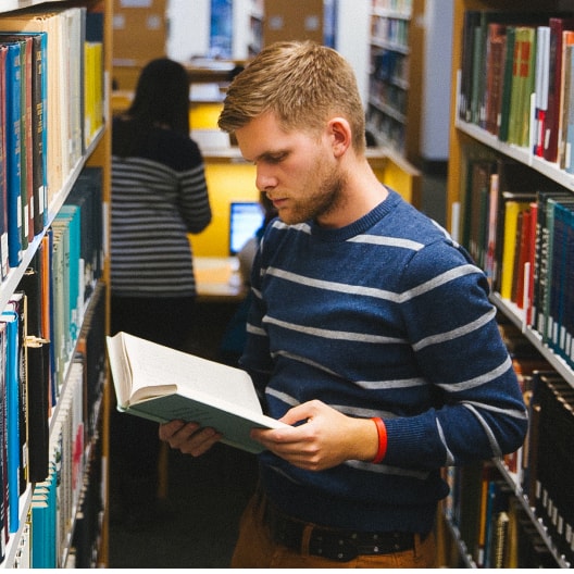 A man in a blue striped sweater reads a book while standing between bookshelves in a library. Another person is working at a desk in the background.