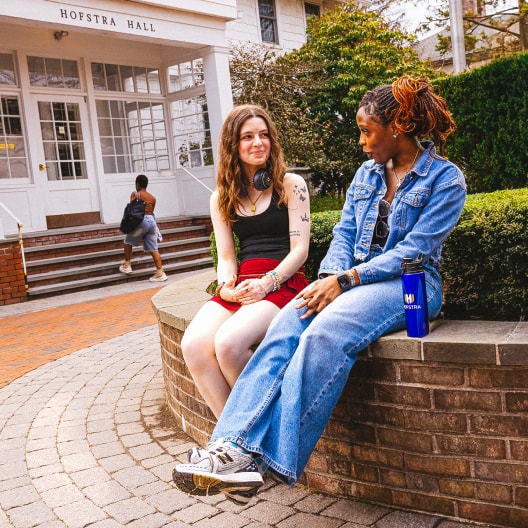 Two students sit and talk on a curved brick wall outside Hofstra Hall. One wears a denim outfit and sneakers, while the other wears a black tank top and red skirt. Trees and another student are in the background.