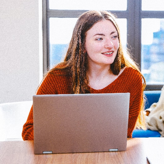 A young woman with long brown hair, wearing an orange sweater, sits at a table with an open laptop, smiling and looking slightly to the side in a brightly lit room.
