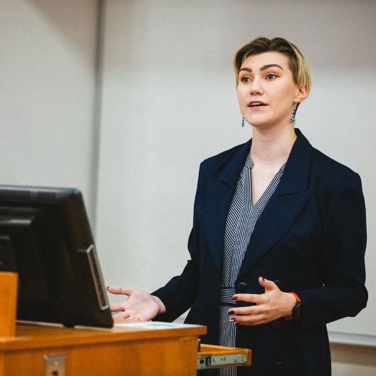 A person in a navy blazer stands at a podium, speaking and gesturing with their hands during a presentation in a classroom or lecture hall.