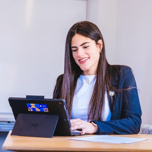 A woman with long brown hair, wearing a blazer, sits at a desk smiling and typing on a tablet with a keyboard in a bright office setting. Papers are scattered on the desk.