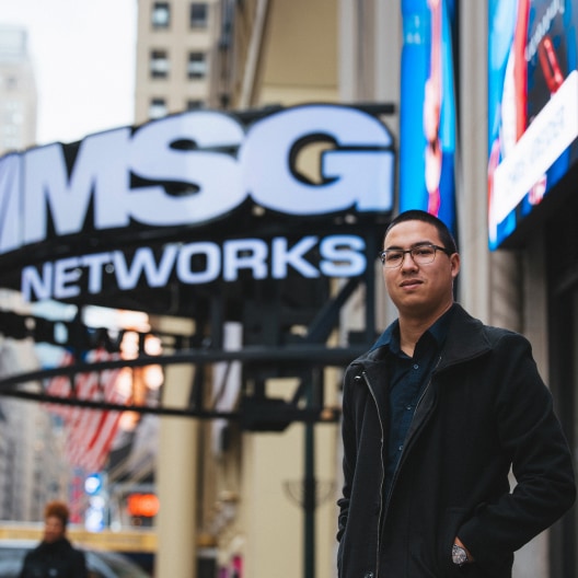 A man wearing glasses and a black coat stands outside near a large MSG Networks sign in an urban setting with tall buildings in the background.