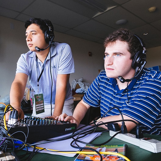 Two men wearing headsets work at an audio mixing console. One stands and adjusts controls, while the other sits and looks ahead, focused. They are in a brightly lit room with papers, wires, and equipment on the table.