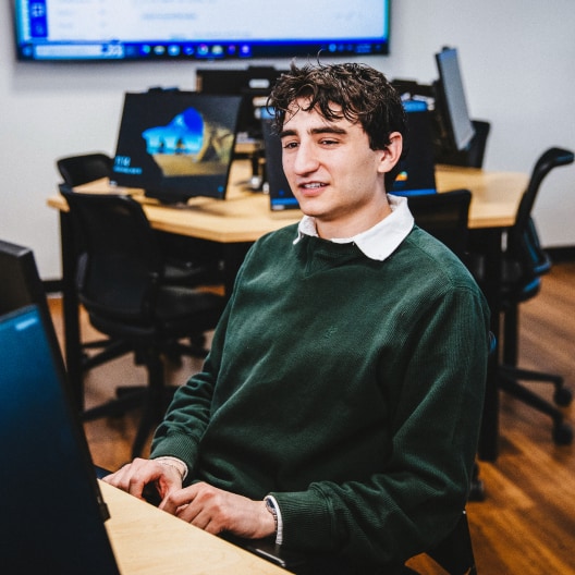 A young man with curly brown hair wearing a green sweater sits at a computer desk in a classroom with several computers and empty chairs around him.