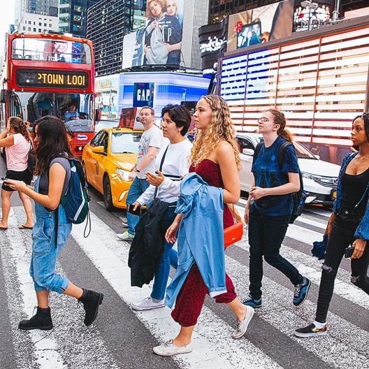 A diverse group of people cross a busy street in a city with bright billboards, a yellow taxi, and a red double-decker tour bus in the background.