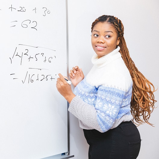 A woman with long braids wearing a white and blue sweater stands by a whiteboard, smiling and holding a marker. Mathematical equations are written on the board.