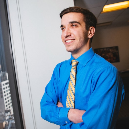 A young man in a bright blue shirt and striped yellow tie stands by a window with his arms crossed, smiling and looking confidently outside. Office buildings and part of the workspace are visible in the background.