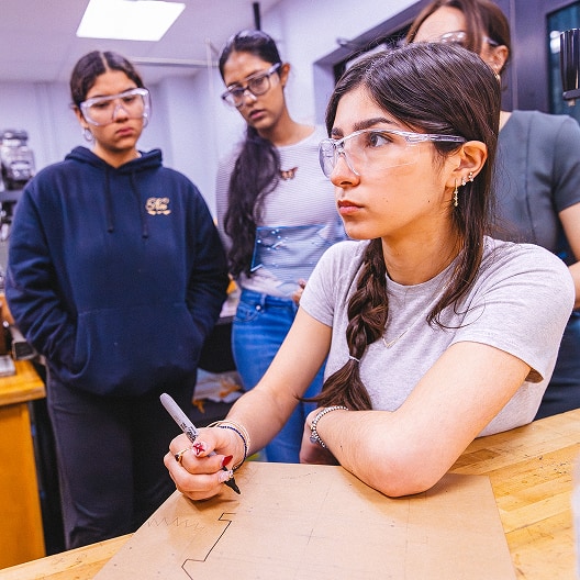 A young woman wearing safety glasses draws on a piece of material at a workbench, while three other women stand nearby observing attentively in a workshop or classroom setting.