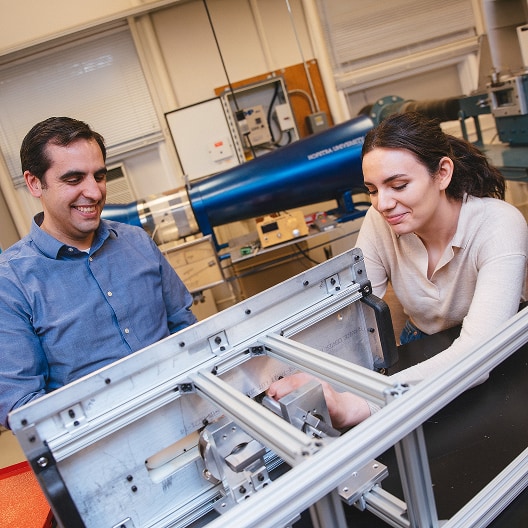 Two people work together on a metal engineering project in a laboratory, surrounded by technical equipment and machinery. One person is smiling while the other adjusts a component inside the frame.