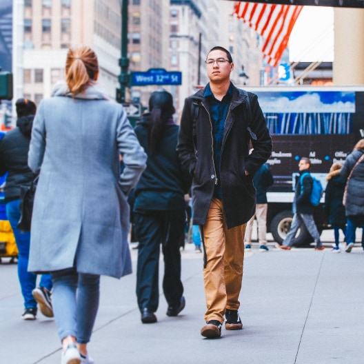 A man in a black coat and tan pants walks down a busy city sidewalk, passing a woman in a gray coat. Other pedestrians and a large American flag are visible in the background.