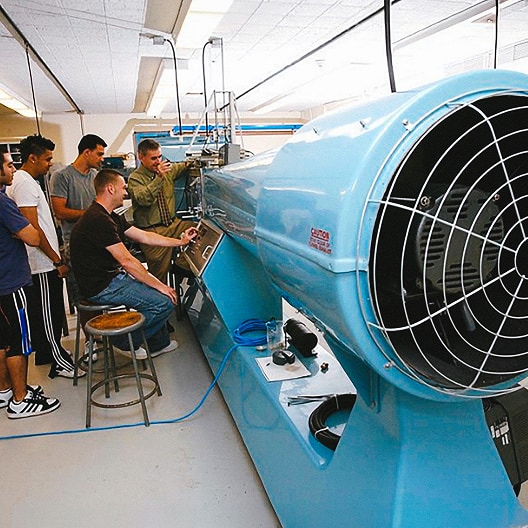 A group of people observe as one person operates a large blue wind tunnel machine in a laboratory setting, with equipment and ceiling lights visible in the background.