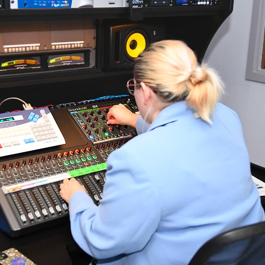 A person with blonde hair in a blue jacket operates an audio mixing console in a recording studio, adjusting knobs and faders surrounded by equipment and monitors.