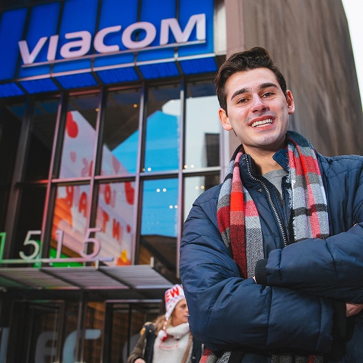 A young man in a winter jacket and plaid scarf stands smiling with arms crossed outside a building with a large blue Viacom sign above the entrance.