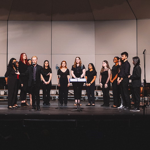 A group of people dressed in black stand on a stage in a semicircle around music stands, appearing to be part of a choir or vocal performance. The background is a simple, light-colored wall.