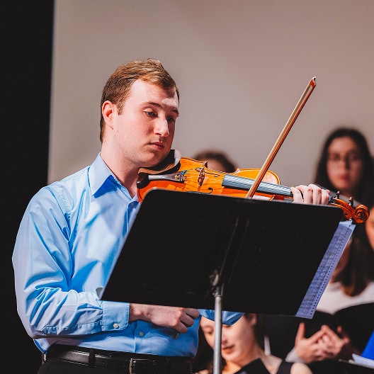 A young man in a blue shirt plays a violin while reading sheet music from a stand. Several people are visible in the blurred background, suggesting a performance or rehearsal setting.