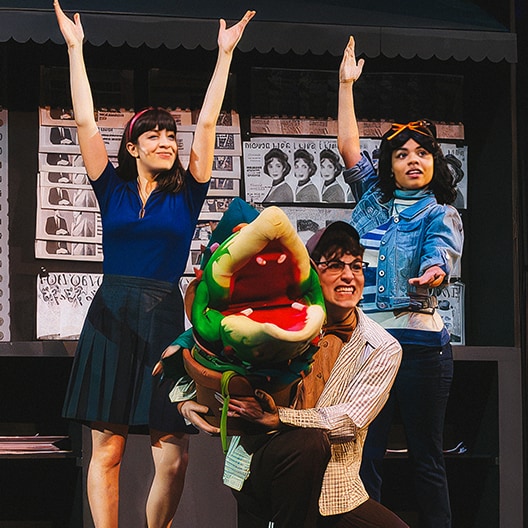 Three actors pose onstage with their arms raised, while one actor kneels holding a large green puppet resembling a carnivorous plant. The background features a wall covered in black-and-white photos.
