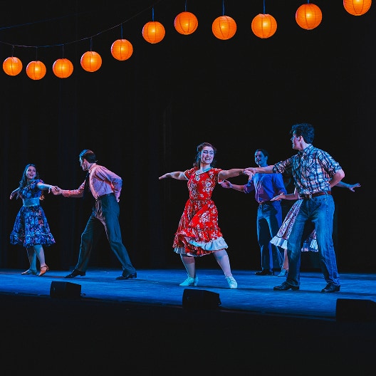 Six people in colorful, retro-style clothing dance in pairs on a stage under a row of glowing orange lanterns, creating a lively and festive atmosphere.